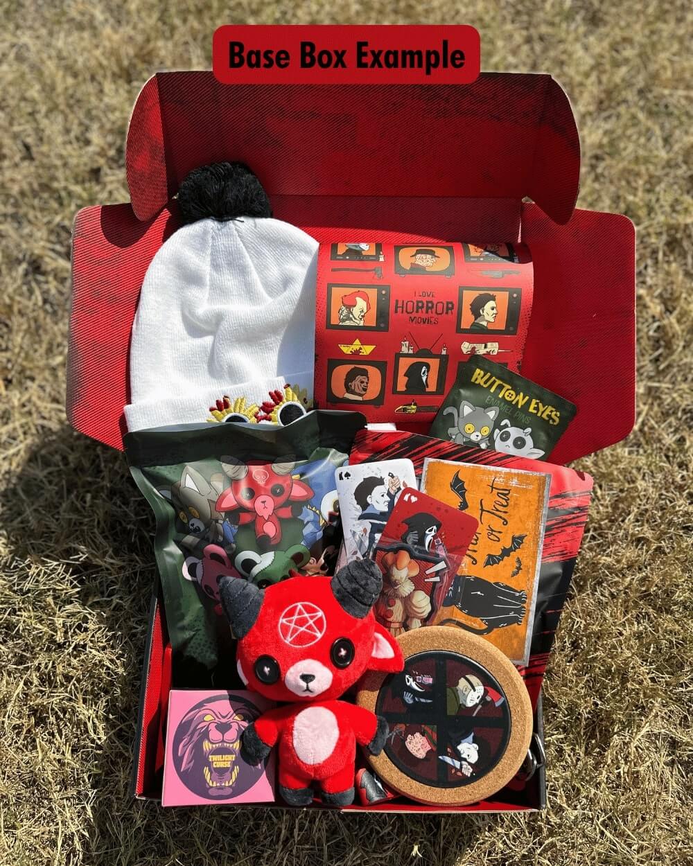 Open red horror box containing various items including a plush toy, cards, plushie blind bag, horror beanie, coaster, and candle on a grassy background.