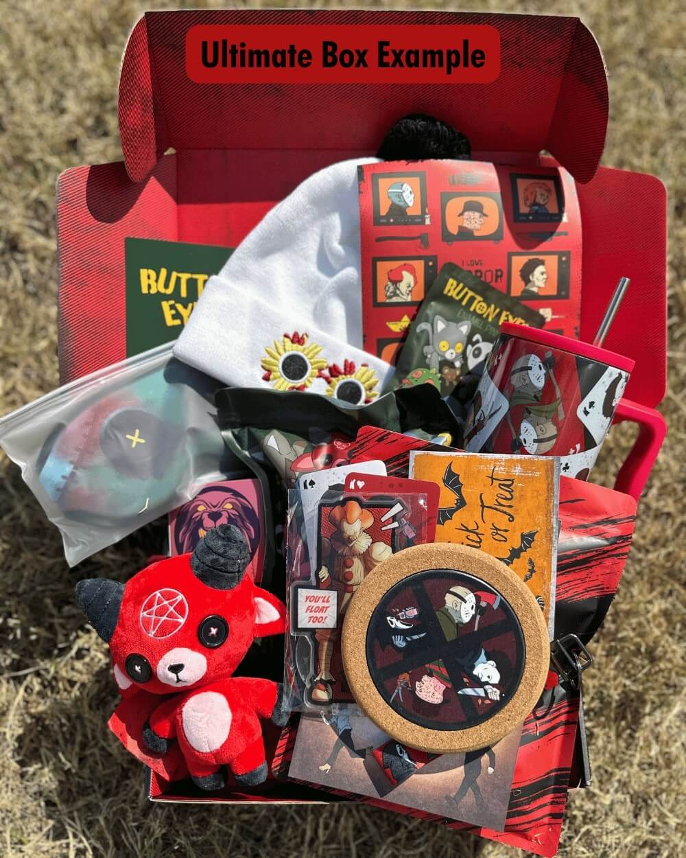 Collection of Halloween-themed horror items including a plush toy, cards, tumbler, sleeping mask, and a hat in a red box labeled 'Ultimate Box Example'.