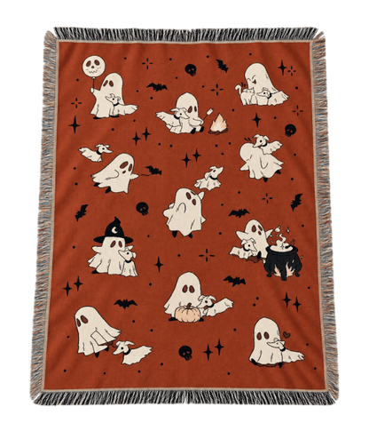 A rectangular woven throw blanket with fringe on all sides, featuring a Halloween pattern of cartoon ghosts, bats, skulls, stars, and a cauldron on a rust-orange background.